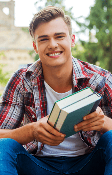 A man in a red, white, and blue flannel holds two books. He is sitting outside, smiling, and looking directly at the camera