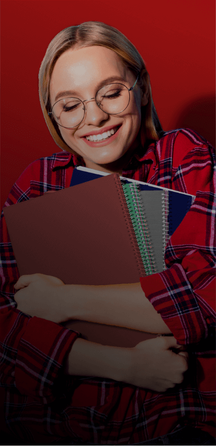 A woman with blonde hair and glasses smiles while holding four multi-colored notebooks to her chest and standing in front of a red background. She has her eyes closed and is wearing a red flannel with white and blue stripes.