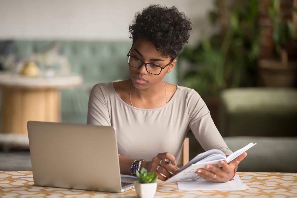 Focused African Student Looking At Laptop Holding Book Doing Research