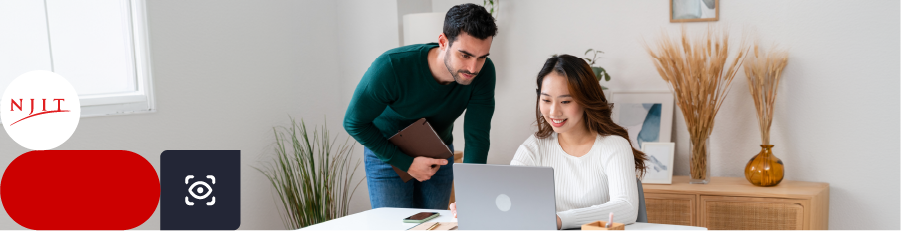 Two people in a modern office, one sitting at a desk with a laptop and the other standing, leaning over with a notebook